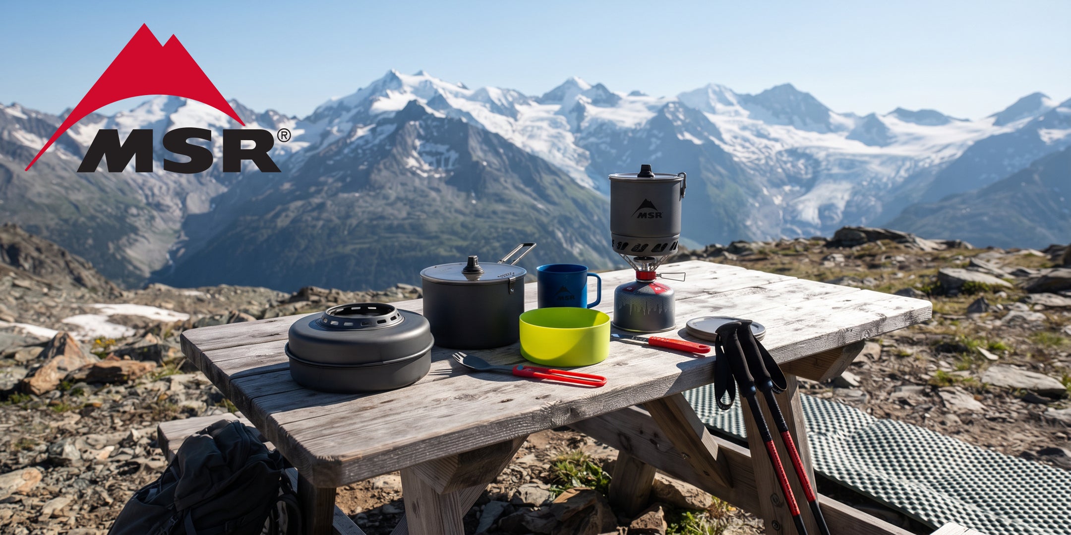 MSR WindBurner e pentole da trekking set fornello da campeggio antivento pentola stoviglie e posate su tavolo da picnic in legno con vista panoramica sulle montagne alpine logo MSR in alto a sinistra