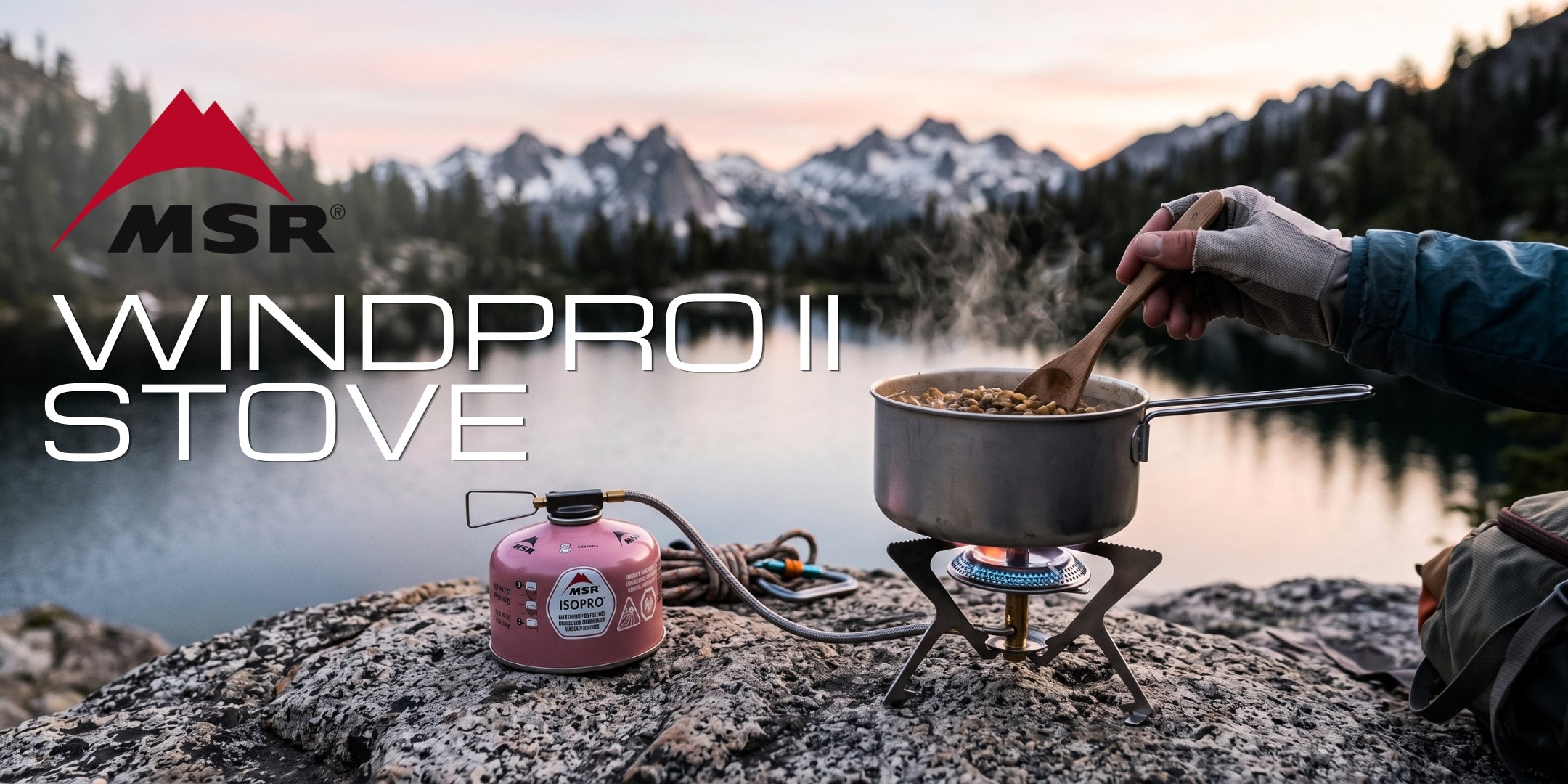 MSR WindPro II fornello a gas con sistema a cartuccia remota, in funzione con pentola per la cottura di cibo su roccia. Contesto outdoor alpino con lago e montagne.