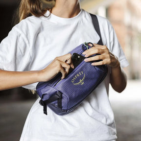 Osprey Transporter Waist Pack marsupio da 3 litri realizzato in tessuto tecnico con trama a griglia e logo frontale, dotato di tasca esterna con zip, cinghie di compressione laterali e cintura regolabile con fibbia a scatto per escursionismo e tempo libero.